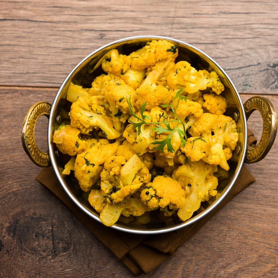 A brass karahi bowl filled with yellow spiced cauliflower florets, garnished with fresh cilantro, on a wooden table.