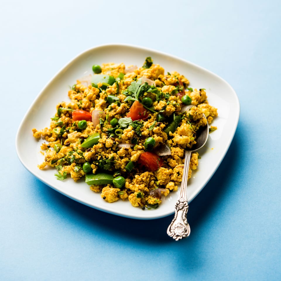 Scrambled paneer bhurji with peas, tomatoes, onions, and cilantro on a white plate with a spoon.